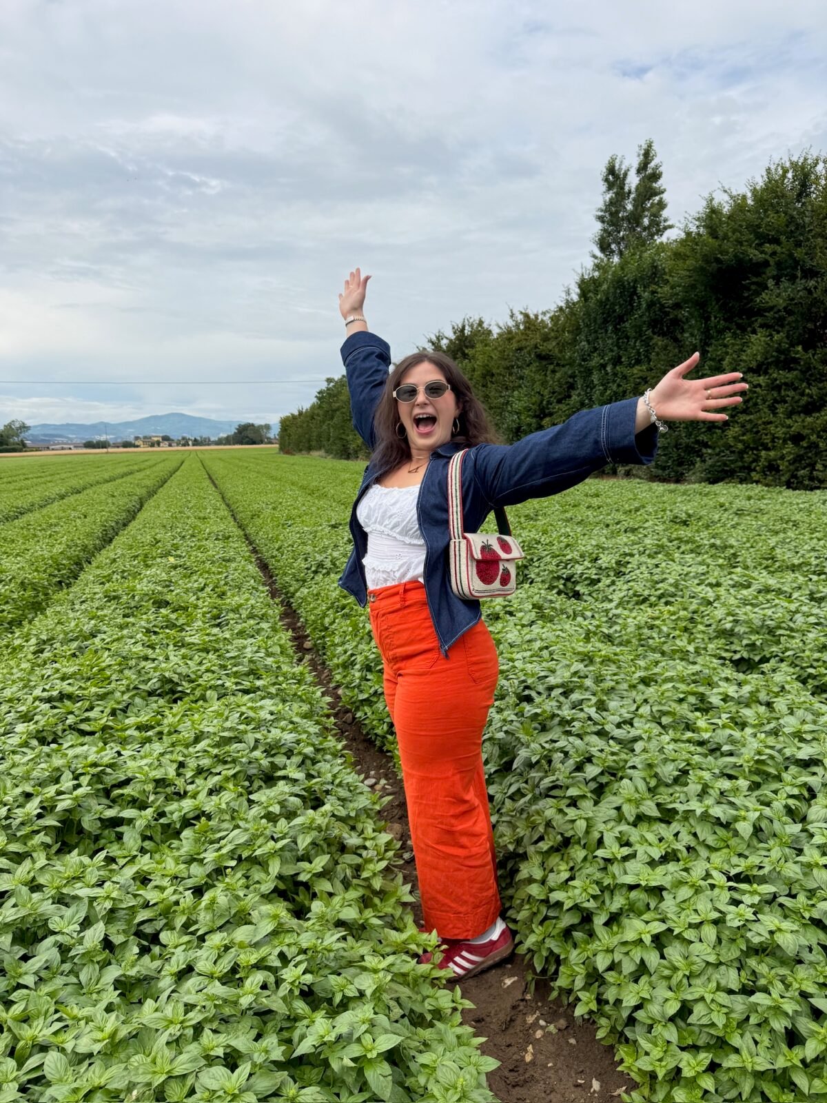 A woman stands in a green field with arms outstretched, smiling excitedly. She wears orange pants, a white top, blue jacket, sunglasses, and a small white purse. Rows of plants and trees stretch into the background under a cloudy sky.