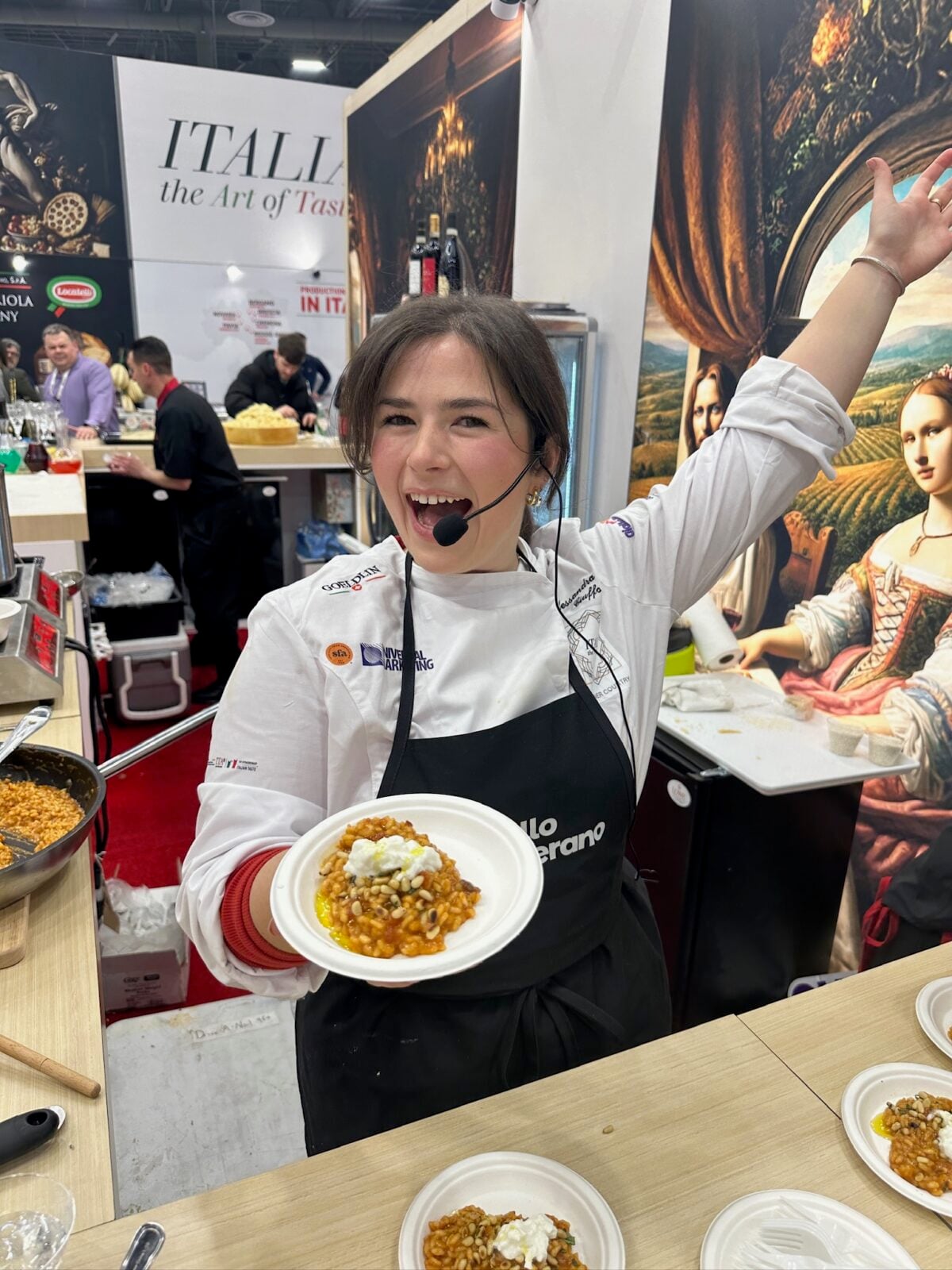 A smiling chef wearing a headset holds up a plate of food in a busy exhibition booth, surrounded by cooking ingredients and banners promoting Italian cuisine.