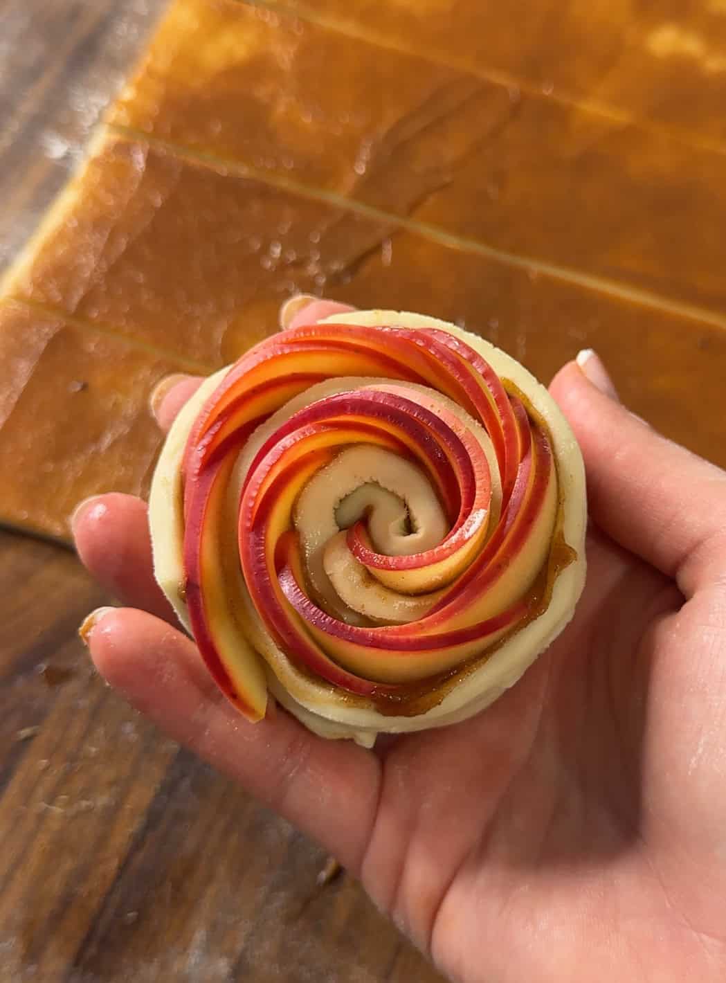 A hand holds a pastry shaped like a rose, made from thin apple slices arranged in a spiral on top of dough. Strips of dough and apple filling are visible on the wooden surface in the background.