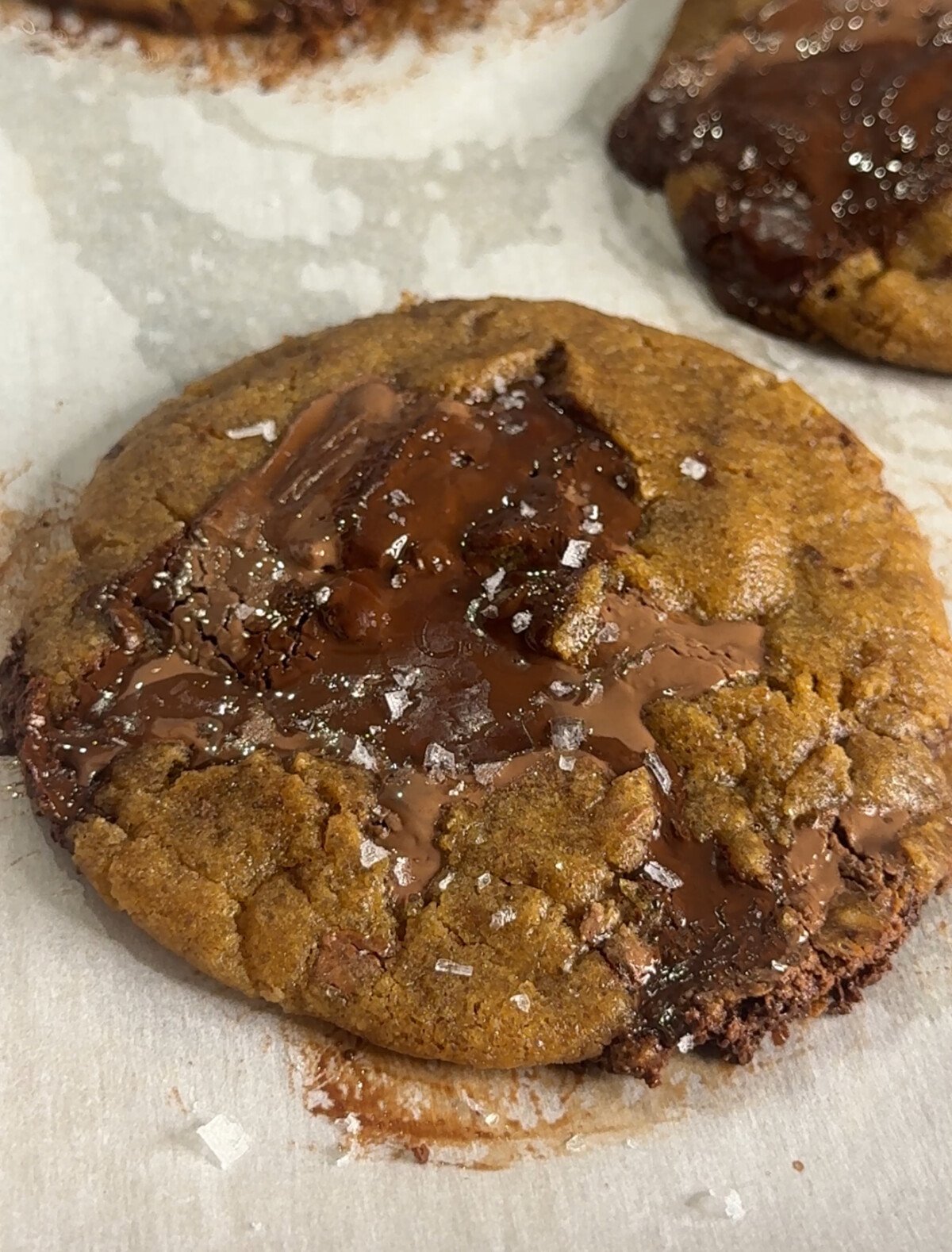A close-up of a freshly baked chocolate chip cookie topped with melted chocolate chunks and a sprinkle of flaky sea salt, resting on parchment paper.