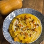 A bowl of creamy butternut squash soup garnished with roasted squash cubes, seeds, herbs, and a drizzle of oil, sits on a wooden surface beside a whole butternut squash.