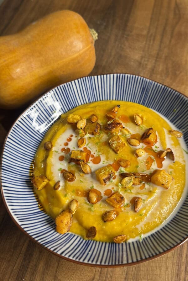 A bowl of creamy butternut squash soup garnished with roasted squash cubes, seeds, herbs, and a drizzle of oil, sits on a wooden surface beside a whole butternut squash.