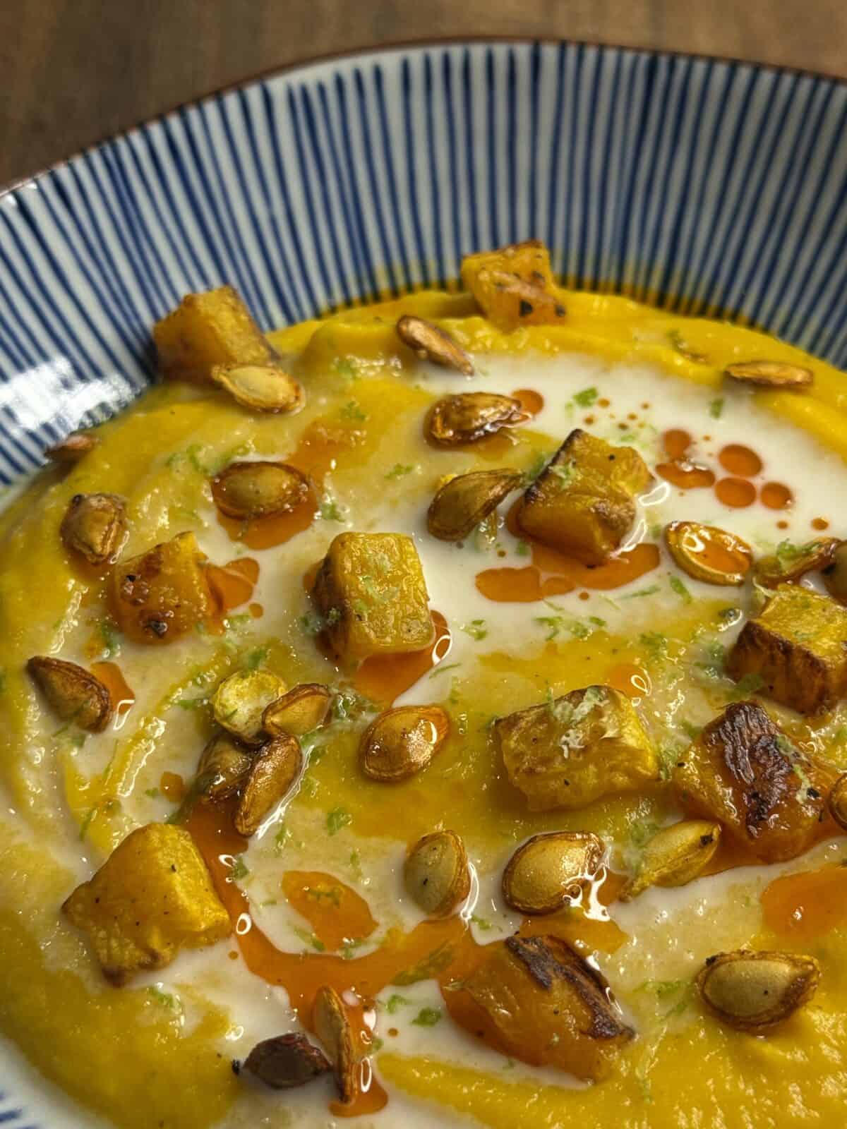 A close-up of creamy pumpkin soup topped with roasted pumpkin cubes, pumpkin seeds, a swirl of cream, and drops of chili oil, served in a striped bowl.