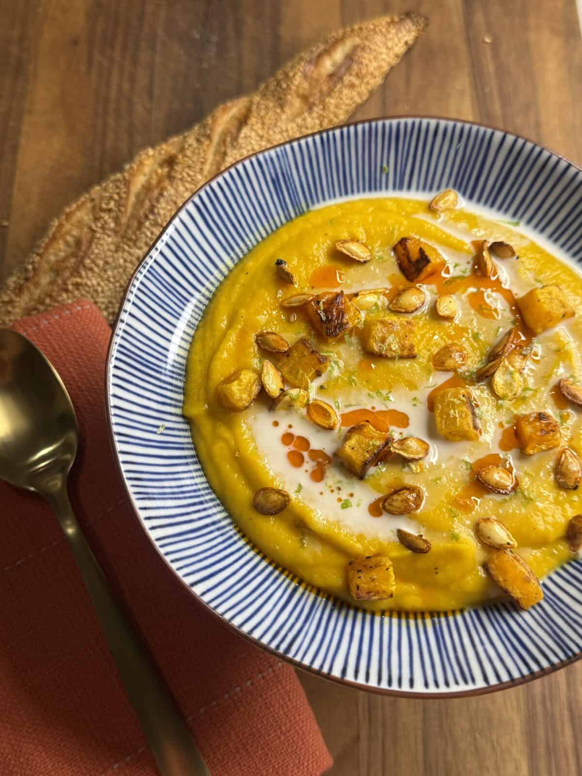 A bowl of creamy orange soup topped with roasted squash cubes, toasted seeds, a drizzle of cream, and red oil, served with a spoon, a red napkin, and a rustic bread slice on a wooden table.