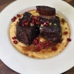 Braised short ribs served on creamy polenta, garnished with pomegranate seeds and lemon zest, presented on a white plate atop a wooden surface.