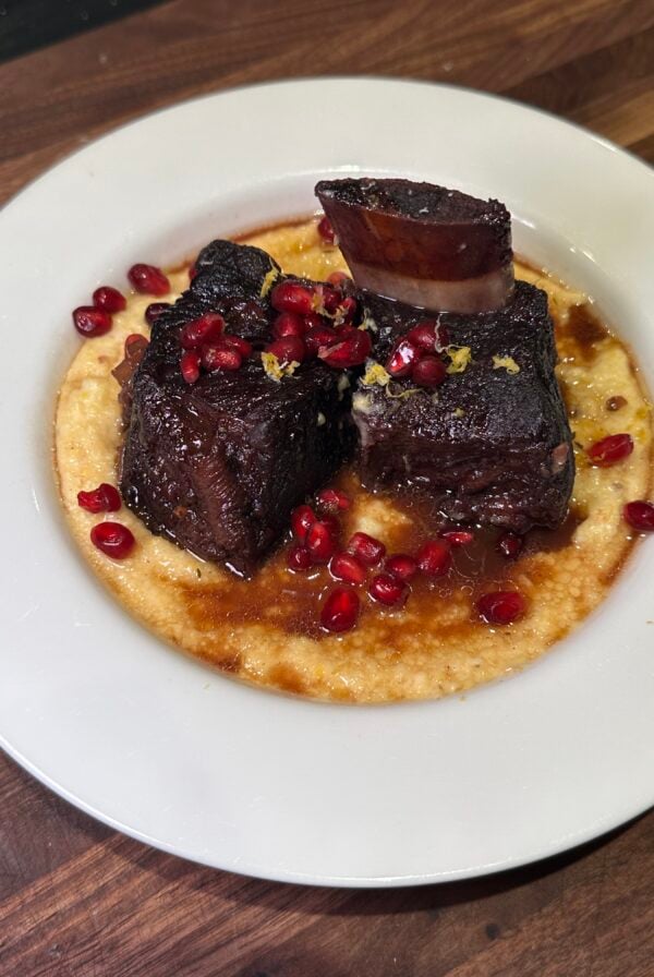 Braised short ribs served on creamy polenta, garnished with pomegranate seeds and lemon zest, presented on a white plate atop a wooden surface.