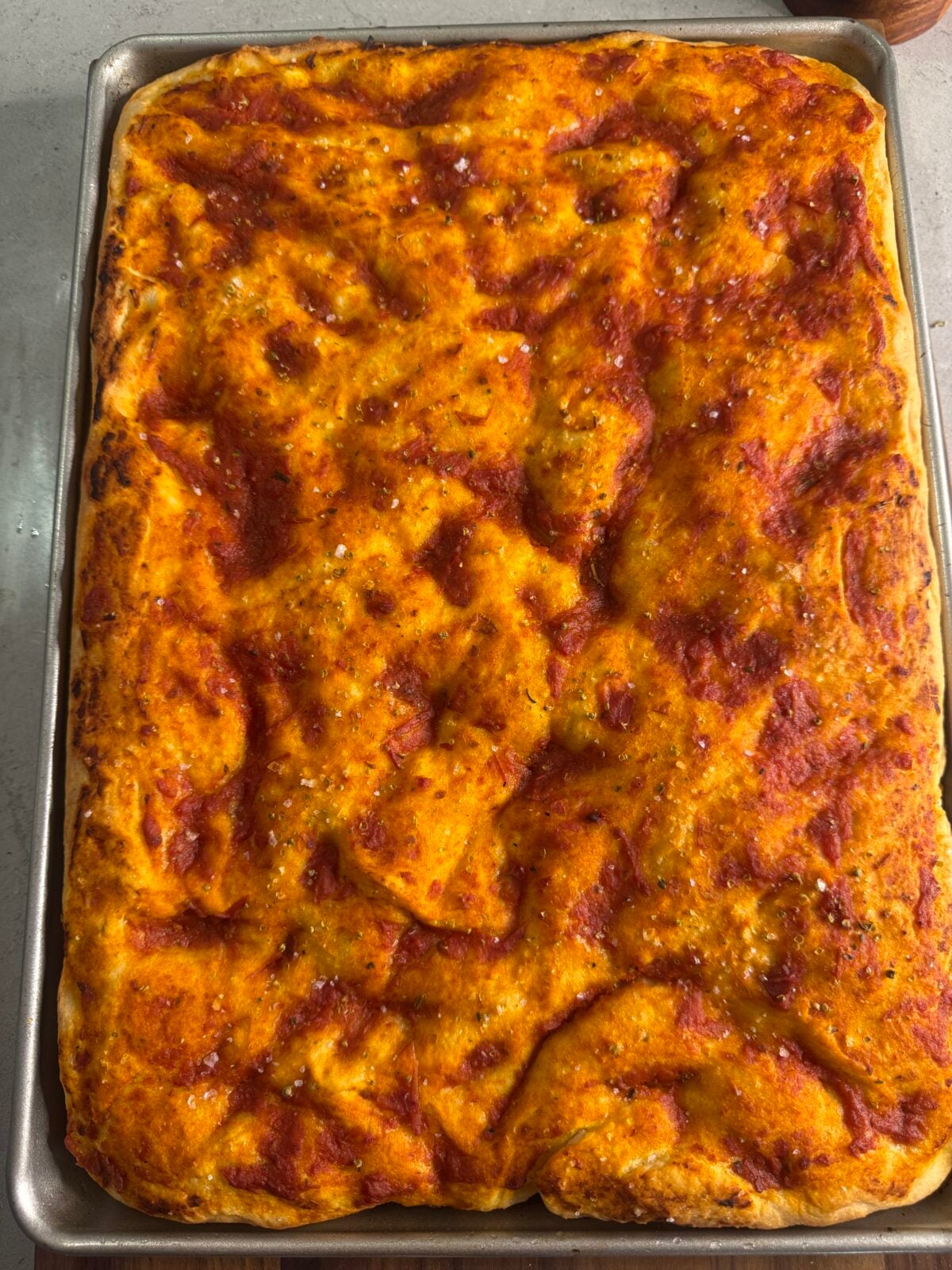 A rectangular sheet pan filled with golden, homemade pizza dough topped with tomato sauce and a sprinkle of coarse salt, fresh out of the oven and resting on a light-colored countertop.