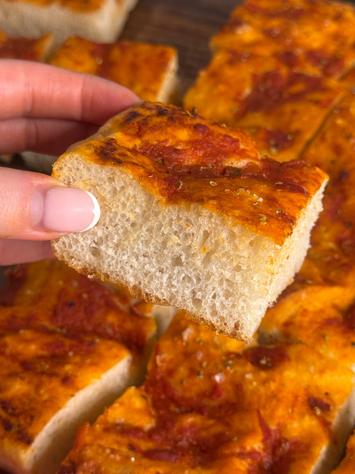 A hand with manicured nails holds a thick, square slice of focaccia bread topped with tomato sauce and herbs. Other pieces of the same focaccia are visible in the background.
