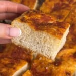 A hand with manicured nails holds a thick, square slice of focaccia bread topped with tomato sauce and herbs. Other pieces of the same focaccia are visible in the background.