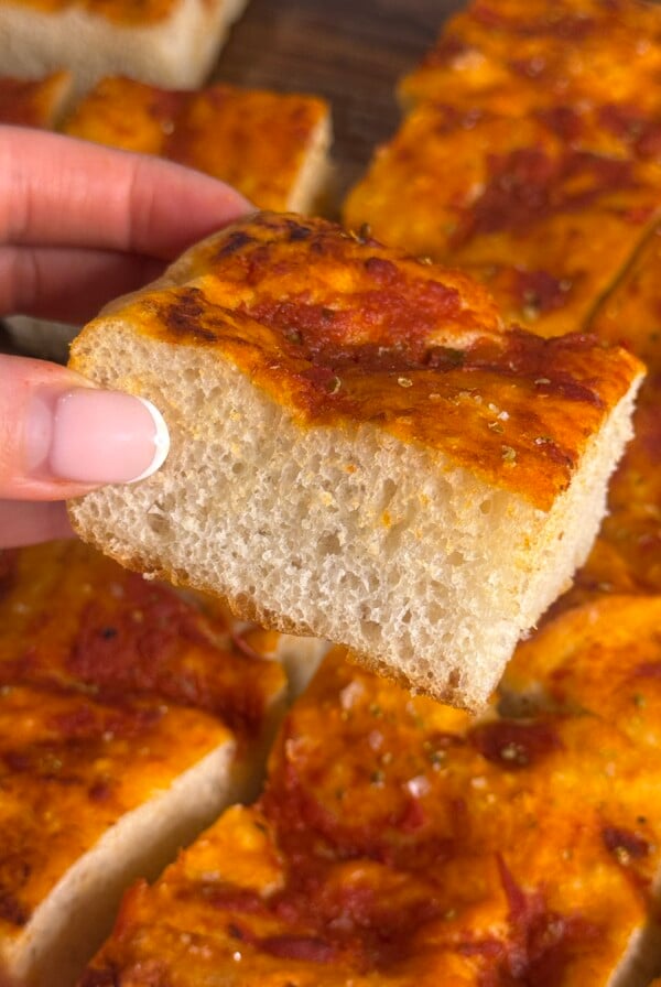 A hand with manicured nails holds a thick, square slice of focaccia bread topped with tomato sauce and herbs. Other pieces of the same focaccia are visible in the background.