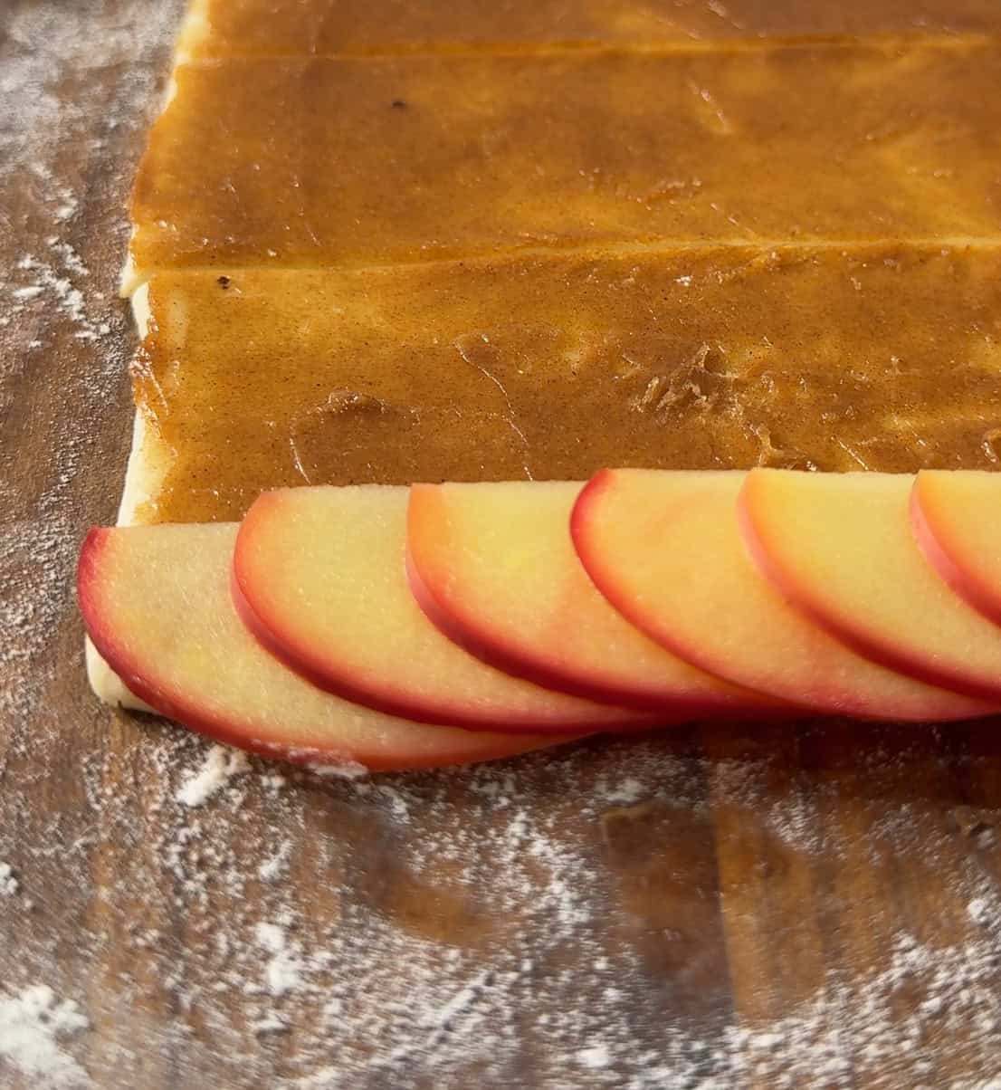 Sliced red apples are neatly arranged on the edge of a rectangular piece of dough spread with brown filling, ready to be rolled on a floured surface.
