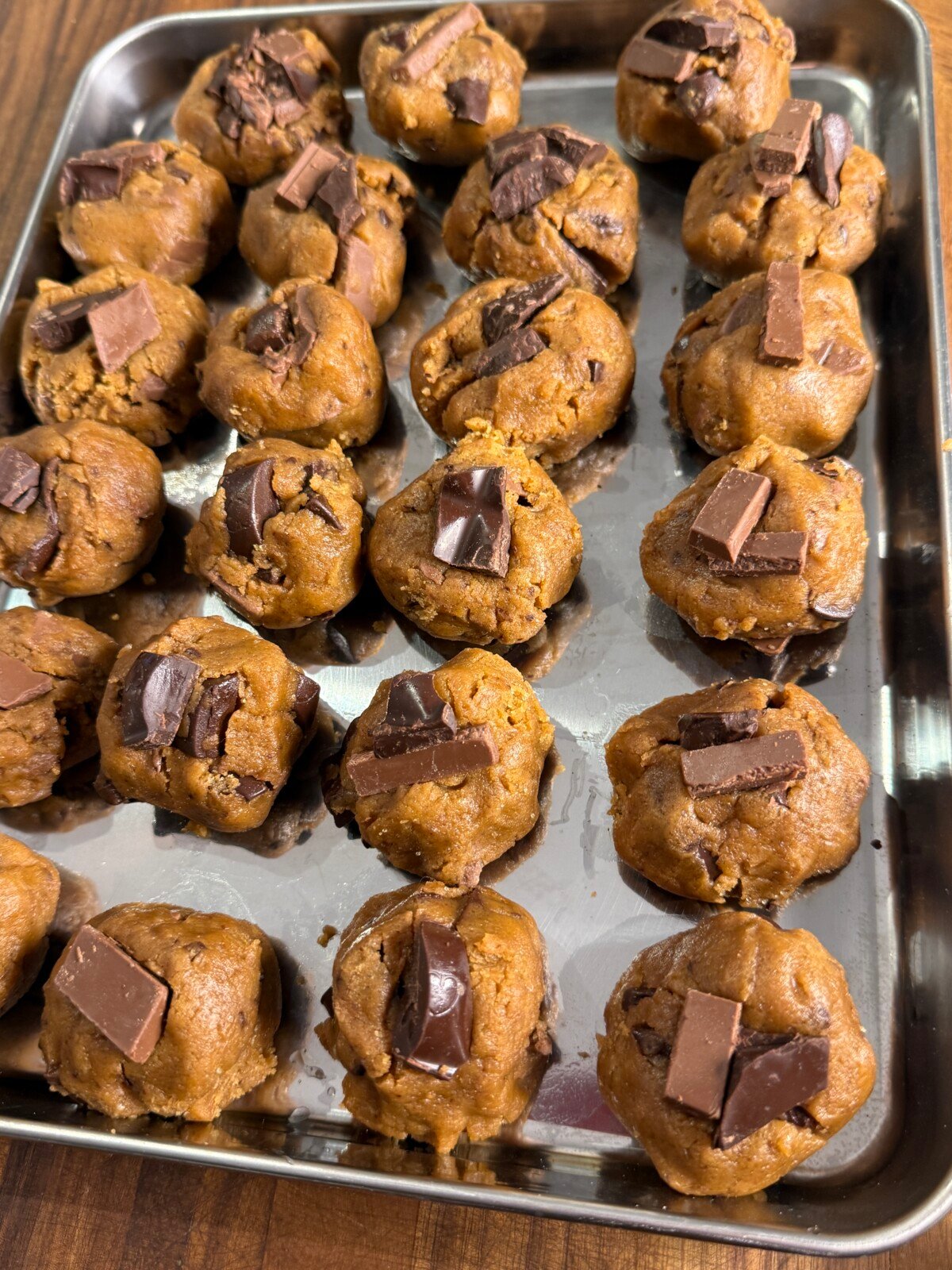 A metal tray with rows of unbaked chocolate chip cookie dough balls, each topped with pieces of chocolate, placed on a wooden surface.
