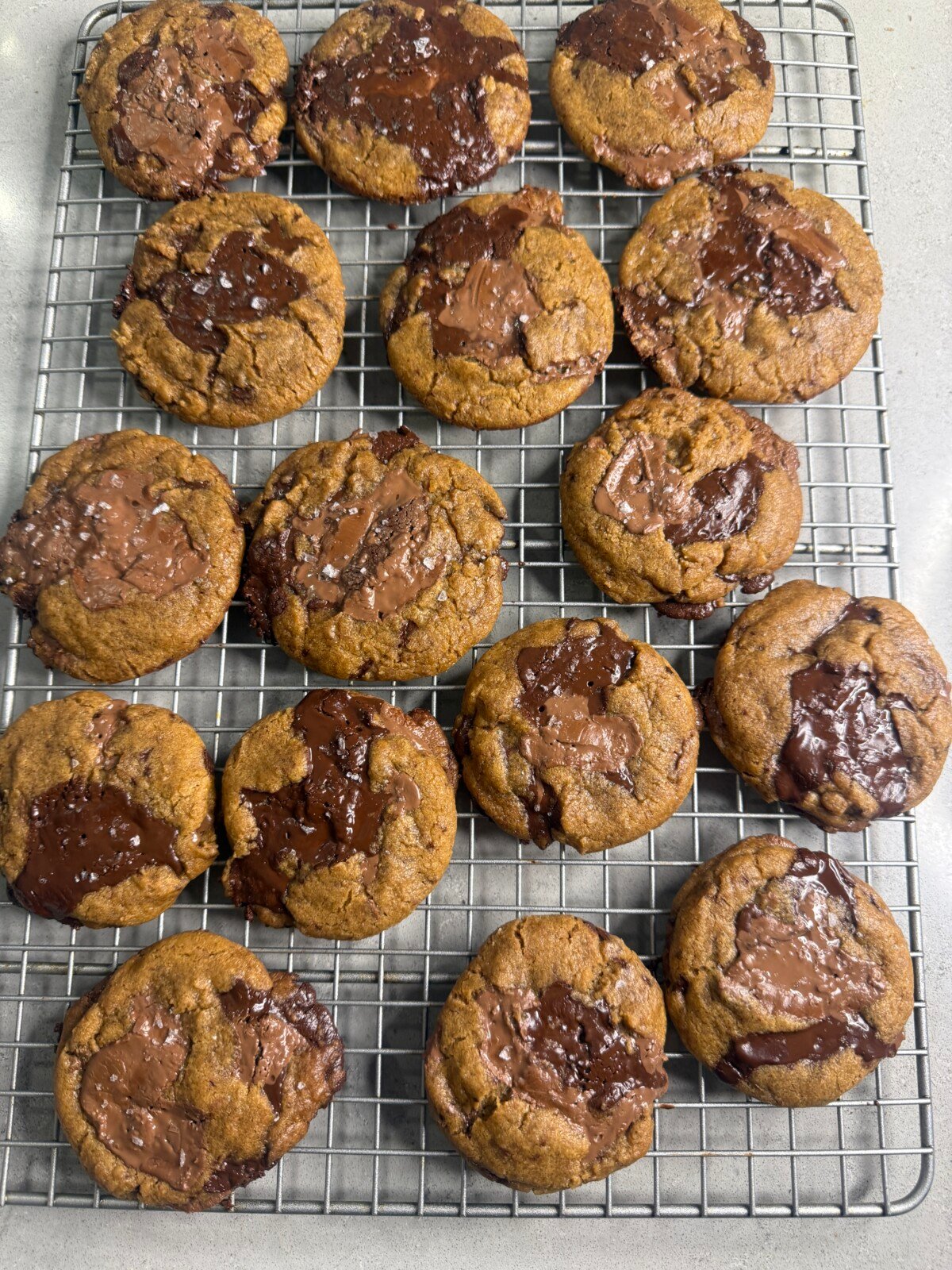 A cooling rack holds 18 freshly baked cookies with melted chocolate swirls on top, some sprinkled with flaky sea salt, arranged in neat rows on a gray countertop.