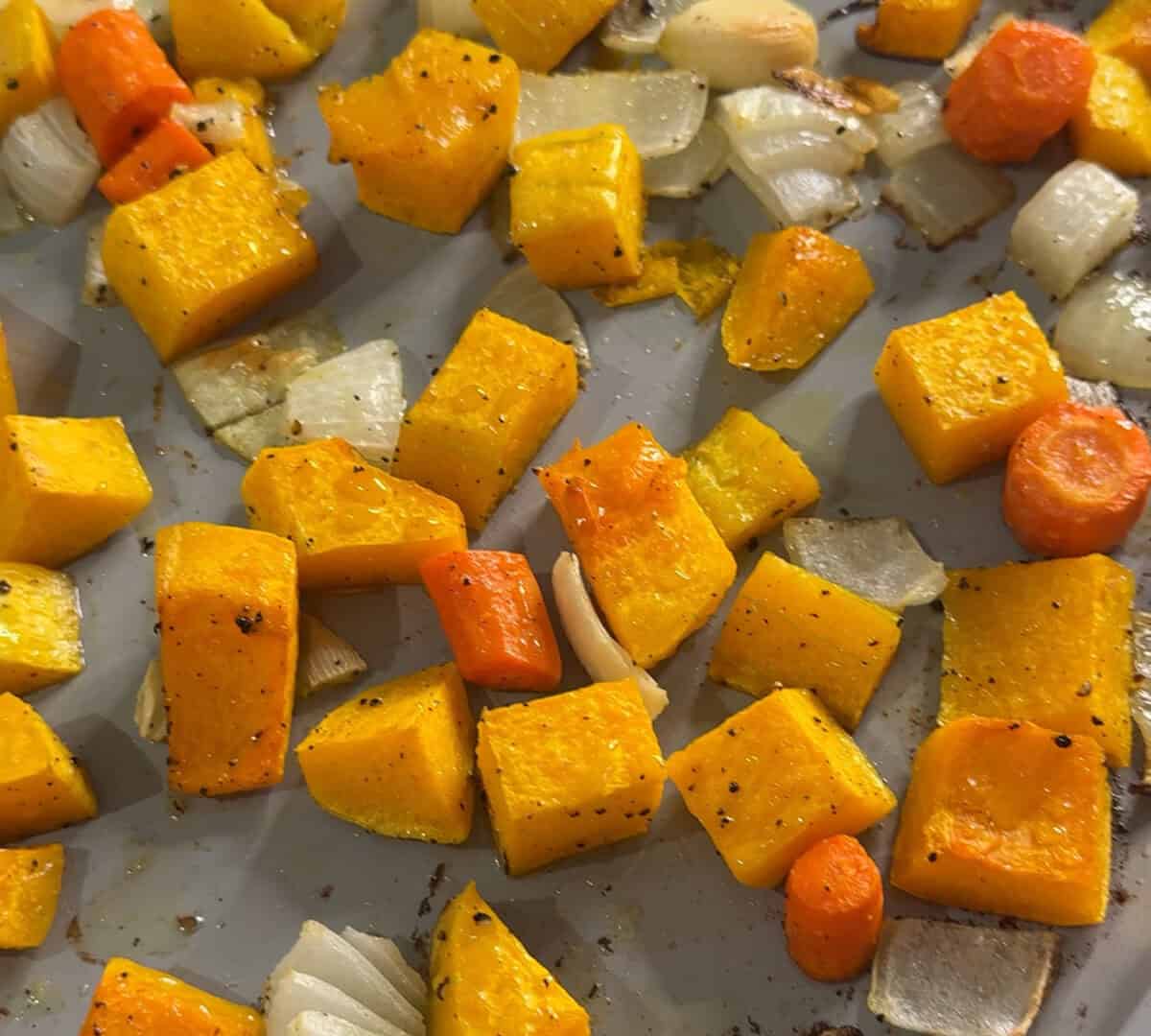 Close-up of roasted vegetables, including cubed butternut squash, carrots, and chopped onions, seasoned with pepper and spread out on a baking sheet.