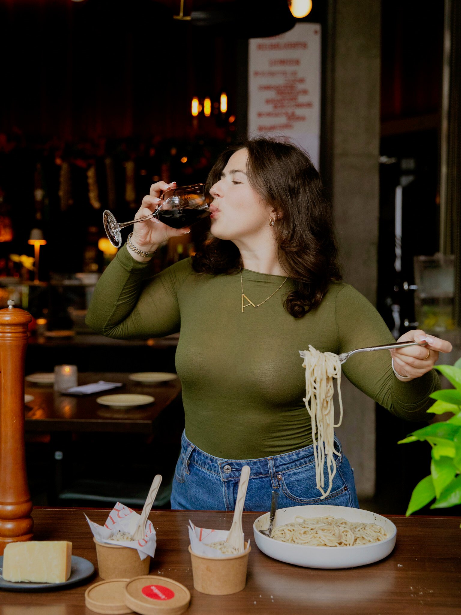 A woman with wavy brown hair wearing a green top and jeans drinks red wine while holding a forkful of pasta over a bowl. She stands at a table with cheese, pasta, and jars, in a warmly lit restaurant.