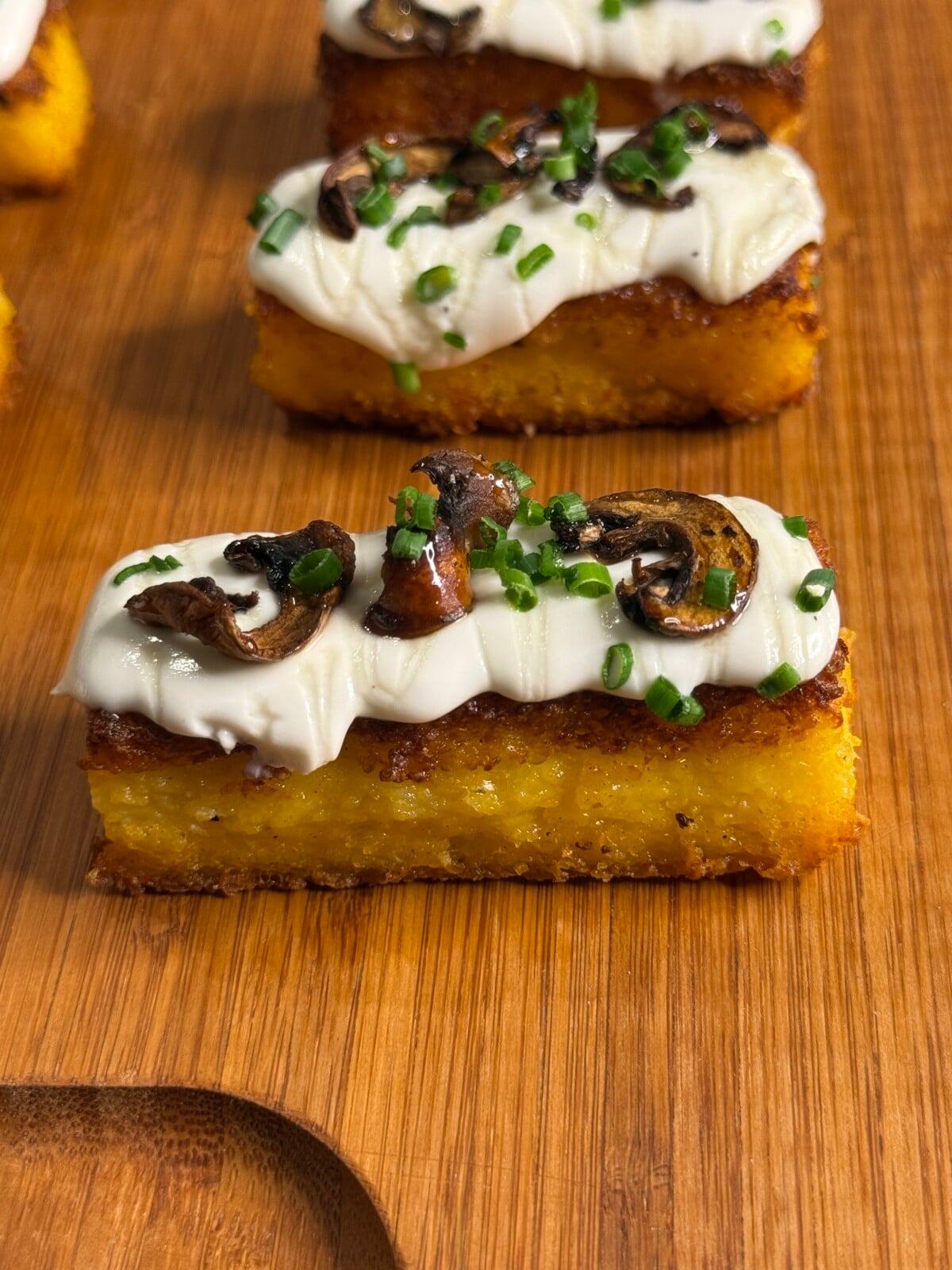 A rectangular slice of golden polenta topped with creamy white sauce, sautéed mushrooms, and chopped chives, served on a wooden board.