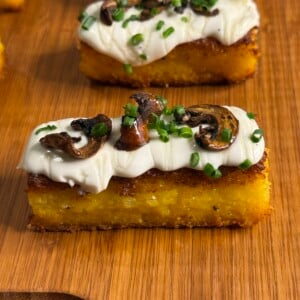 A rectangular slice of golden polenta topped with creamy white sauce, sautéed mushrooms, and chopped chives, served on a wooden board.