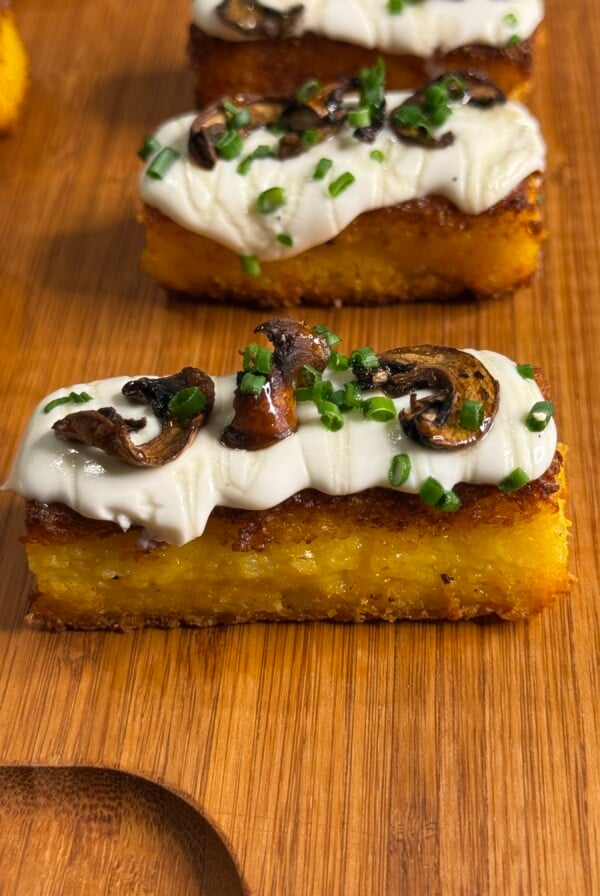 A rectangular slice of golden polenta topped with creamy white sauce, sautéed mushrooms, and chopped chives, served on a wooden board.