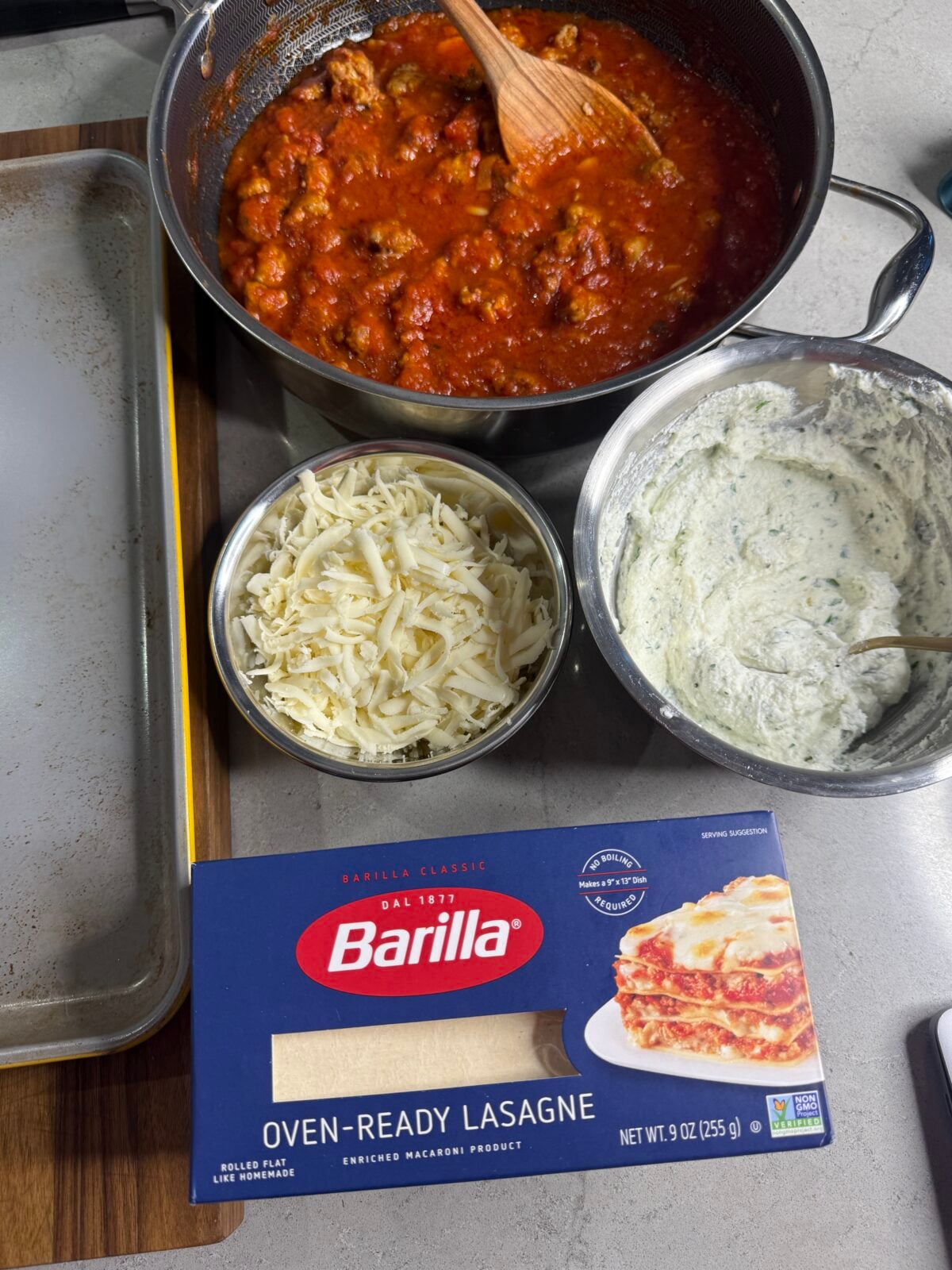 A pot of meat sauce, a bowl of shredded cheese, a bowl of ricotta mixture, and a box of Barilla oven-ready lasagne noodles are arranged on a kitchen counter, ready for lasagna assembly.