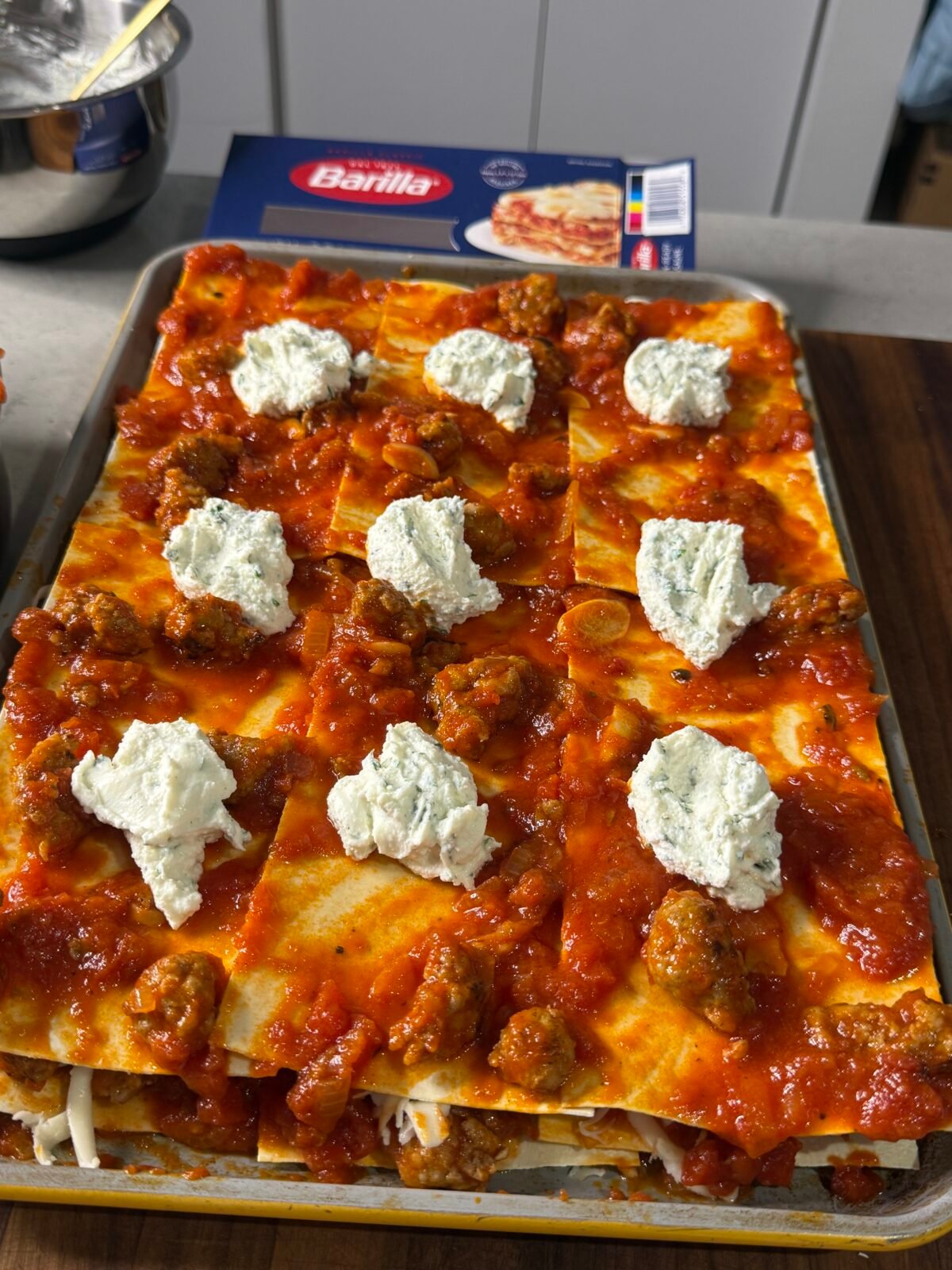 A tray of uncooked lasagna with layers of pasta, tomato sauce, sausage, and dollops of ricotta cheese sits on a kitchen counter. A box of Barilla lasagna noodles is visible in the background.