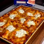 A freshly baked lasagna with golden, melted cheese sits in a baking dish on a wooden surface. A box of Barilla oven-ready lasagne noodles is visible in the background.
