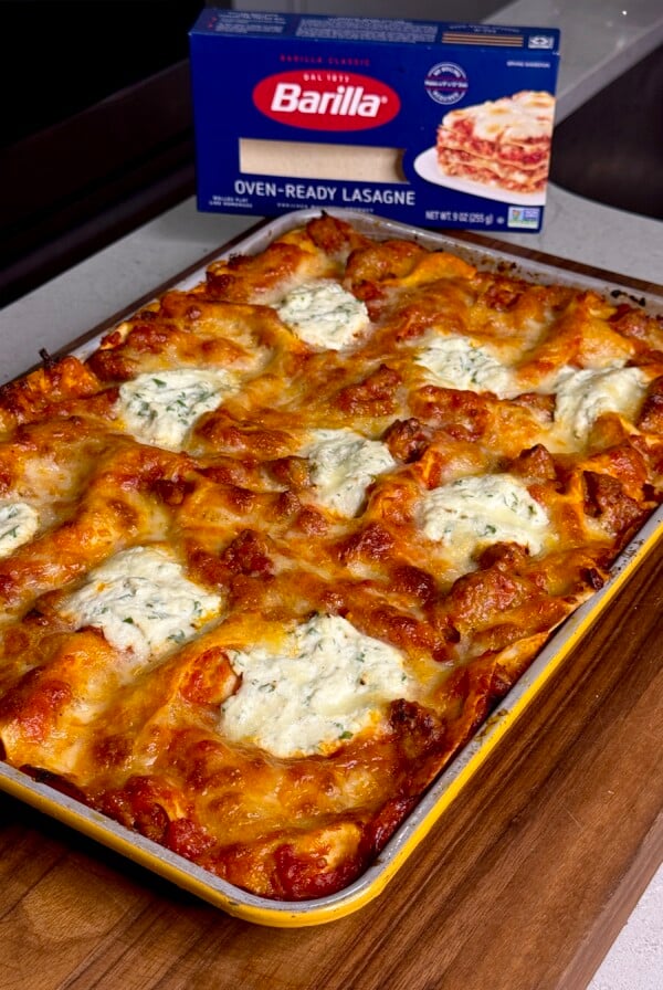 A freshly baked lasagna with golden, melted cheese sits in a baking dish on a wooden surface. A box of Barilla oven-ready lasagne noodles is visible in the background.