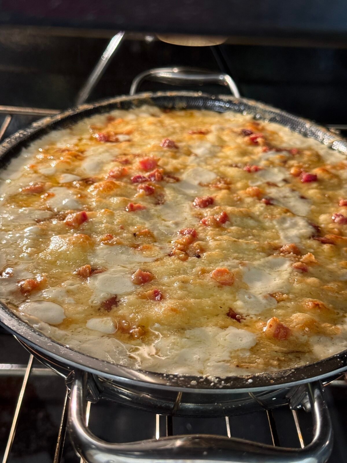 A close-up of a cheesy dish with bubbling melted cheese and pieces of bacon or ham, baking in a round pan inside an oven. The surface appears golden and slightly crispy.