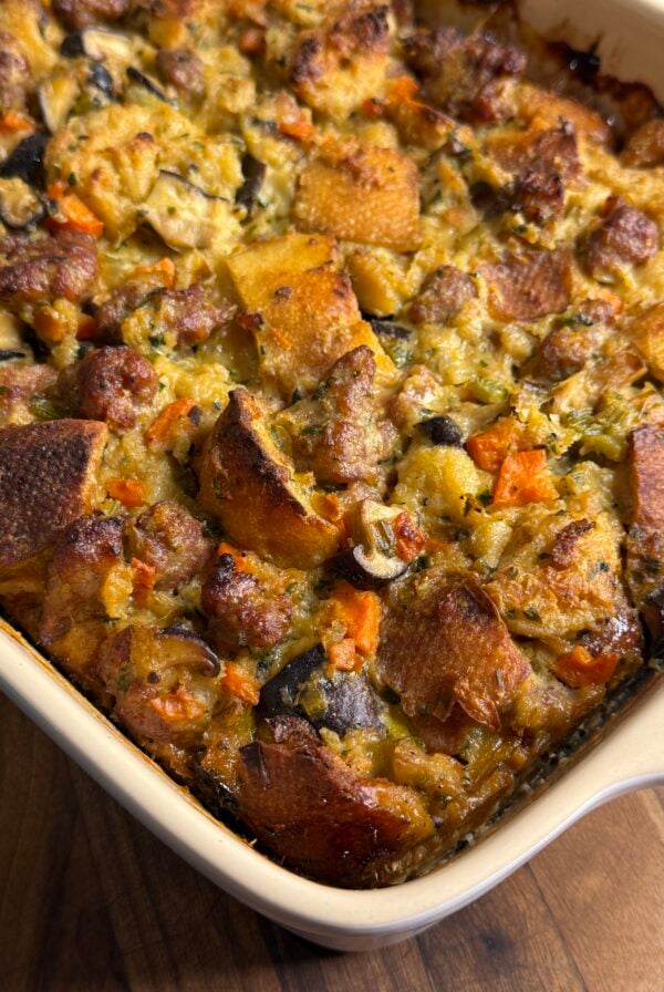 A close-up of a baked casserole dish filled with golden brown stuffing made of bread cubes, sausage, vegetables like carrots and celery, and herbs, resting on a wooden surface.