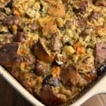 A close-up of a baked casserole dish filled with bread stuffing, featuring browned bread cubes, sausage, carrots, celery, and herbs. The top is golden and crispy.