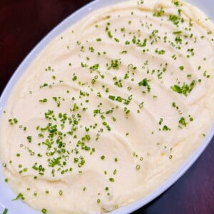 A white oval dish filled with creamy mashed potatoes, topped with finely chopped chives and set on a dark wooden table.