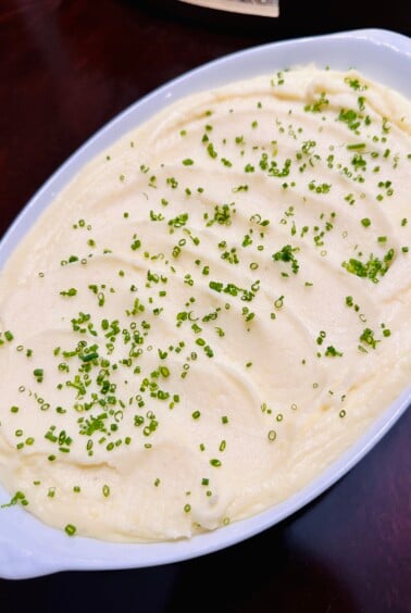 A white oval dish filled with creamy mashed potatoes, topped with finely chopped chives and set on a dark wooden table.