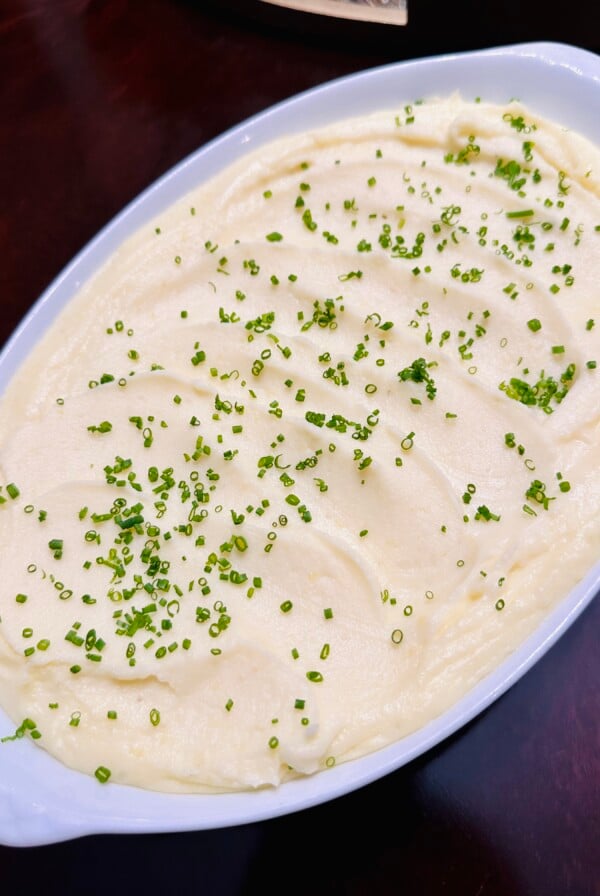 A white oval dish filled with creamy mashed potatoes, topped with finely chopped chives and set on a dark wooden table.