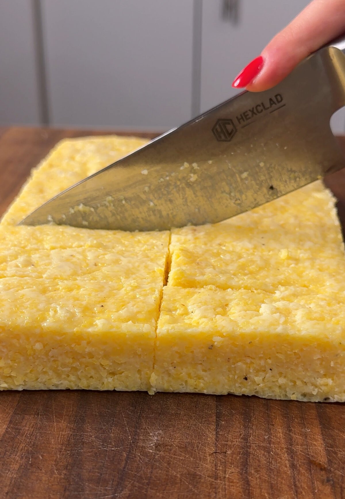 A large knife slices through a thick, rectangular block of yellow polenta on a wooden cutting board. A hand with red-painted nails holds the knife.
