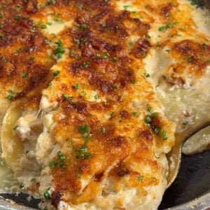 A baked cheesy casserole with a golden-brown crust, garnished with chopped herbs, shown in a skillet with a serving spoon scooping out a portion.