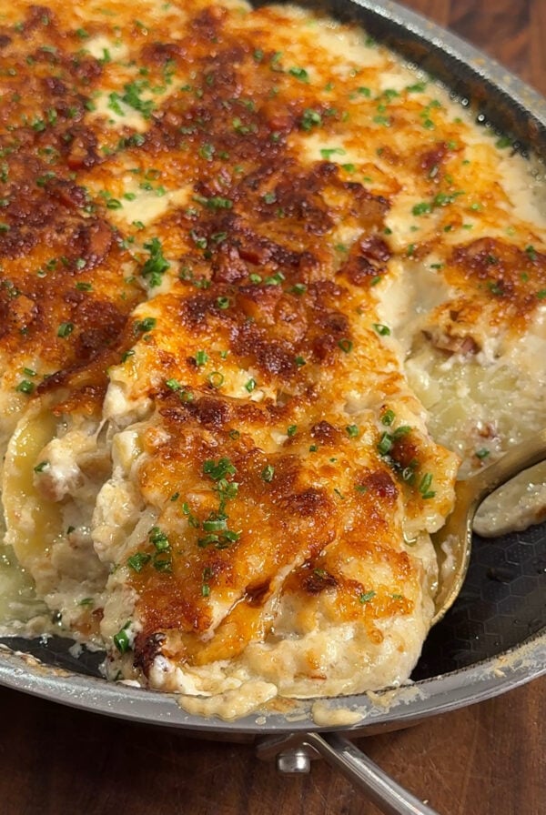 A baked cheesy casserole with a golden-brown crust, garnished with chopped herbs, shown in a skillet with a serving spoon scooping out a portion.