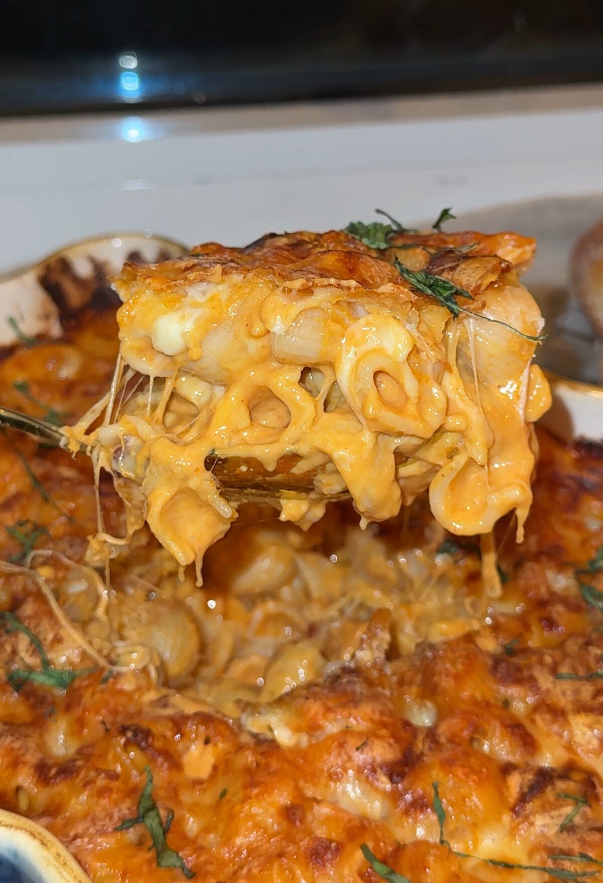 A close-up of a cheesy baked macaroni and cheese being lifted from a casserole dish, with gooey melted cheese stretching between the pasta and the pan.