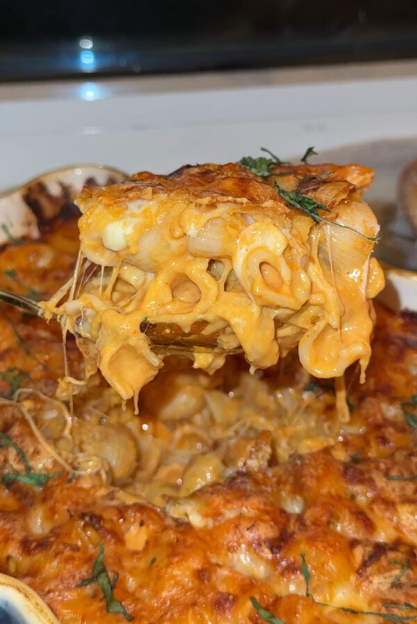 A close-up of a cheesy baked macaroni and cheese being lifted from a casserole dish, with gooey melted cheese stretching between the pasta and the pan.