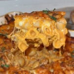 A close-up of a cheesy baked macaroni and cheese being lifted from a casserole dish, with melted cheese stretching and garnished with fresh herbs.