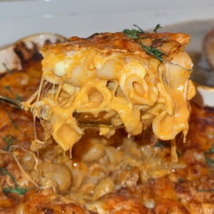 A close-up of a cheesy baked macaroni and cheese being lifted from a casserole dish, with melted cheese stretching and garnished with fresh herbs.