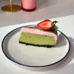 A slice of green matcha cheesecake topped with pink cream and half a fresh strawberry, served on a white plate with a blue rim. A pink drink is visible in the blurred background.
