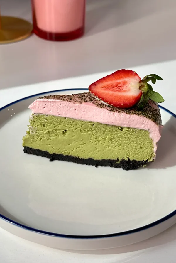 A slice of green matcha cheesecake topped with pink cream and half a fresh strawberry, served on a white plate with a blue rim. A pink drink is visible in the blurred background.