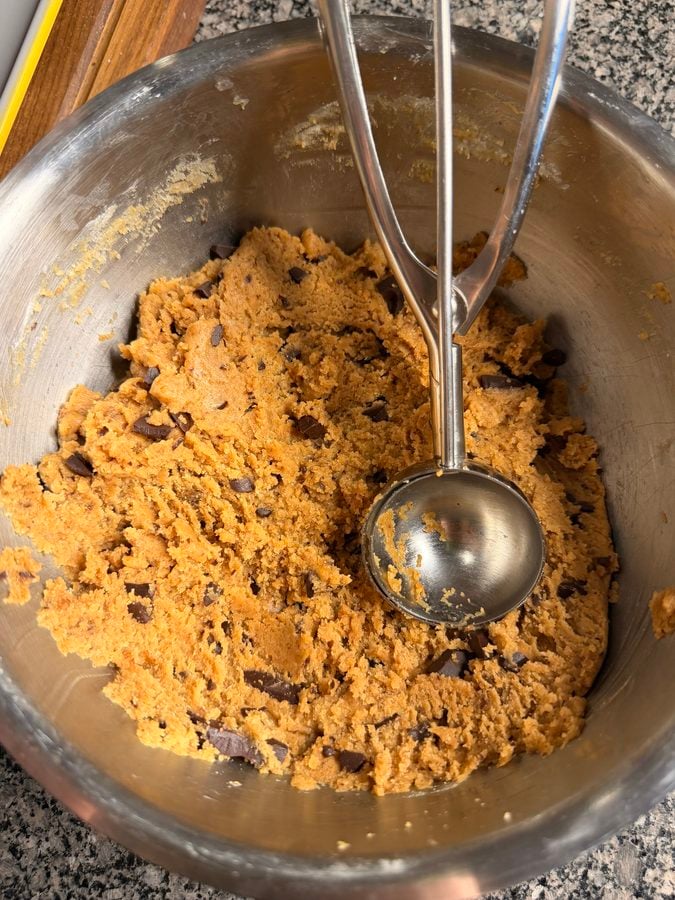 A metal mixing bowl with chocolate chip cookie dough and a metal ice cream scoop resting on top, on a granite countertop.