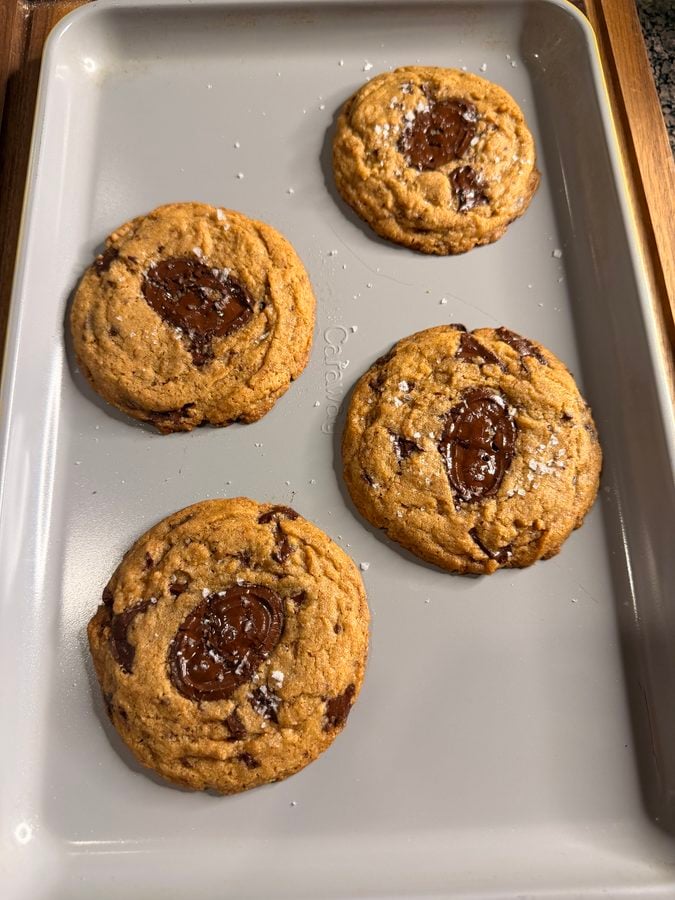 Four chocolate chip cookies with melted chocolate centers and a sprinkle of sea salt sit on a light gray baking sheet.
