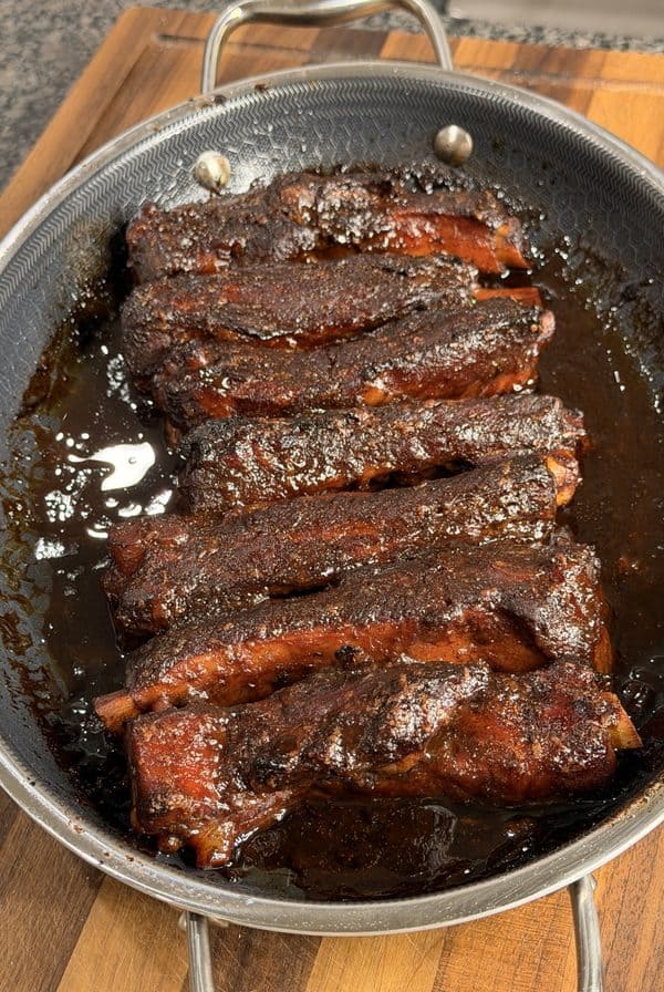 A pan filled with glazed, seasoned pork ribs resting on a wooden cutting board. The ribs are coated in a dark, sticky sauce and appear freshly cooked.