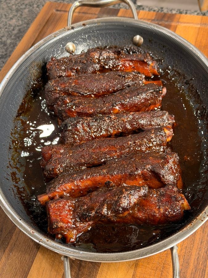 A pan filled with glazed, seasoned pork ribs resting on a wooden cutting board. The ribs are coated in a dark, sticky sauce and appear freshly cooked.