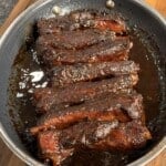 A pan filled with glazed, cooked pork ribs coated in a dark, sticky barbecue sauce sits on a wooden cutting board.