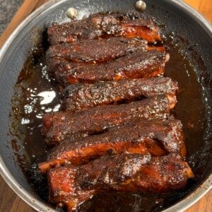 A pan filled with glazed, cooked pork ribs coated in a dark, sticky barbecue sauce sits on a wooden cutting board.