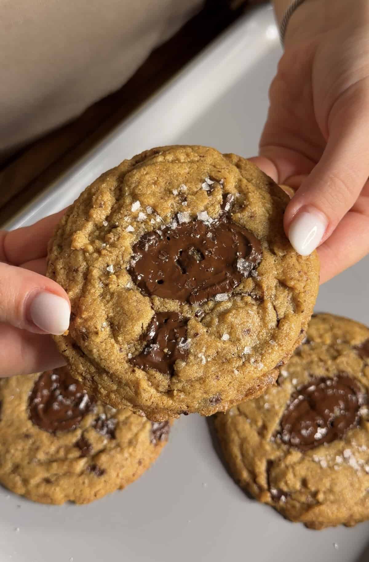 A person holds a chocolate chip cookie topped with sea salt flakes, with two more cookies visible below on a white plate.