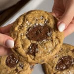 A person holds a chocolate chip cookie topped with sea salt flakes, with two more cookies visible below on a white plate.