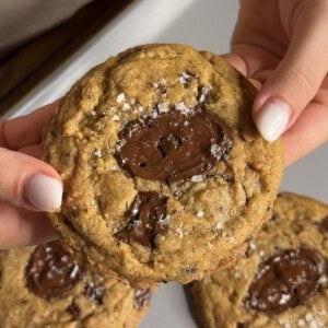 A person holds a chocolate chip cookie topped with sea salt flakes, with two more cookies visible below on a white plate.